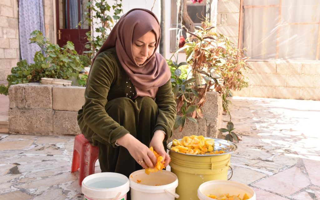 Woman in green dress and mauve hijab sits in front of garden preparing food over white and green buckets.