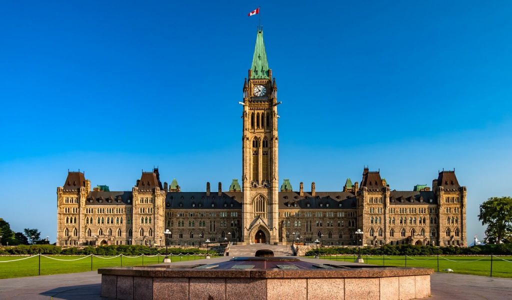 An image of Canada's eternal flame in front of the Parliament of Canada's centre block. The sky is blue, and it is daytime.