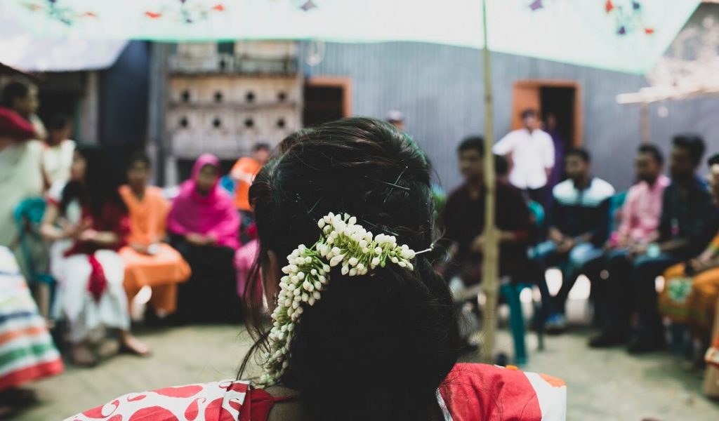 In the foreground, there is a young Bangladeshi woman with white flowers in her hair wearing a red sari. She is taking part in a workshop outdoors, and she is sitting in a circle of at least a dozen other participants.