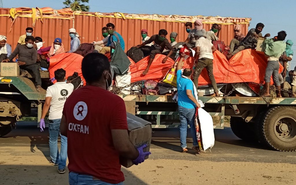 Man in red Oxfam shirt with back turned to camera carrying a box of food and water to migrant works stranded on truck in background.