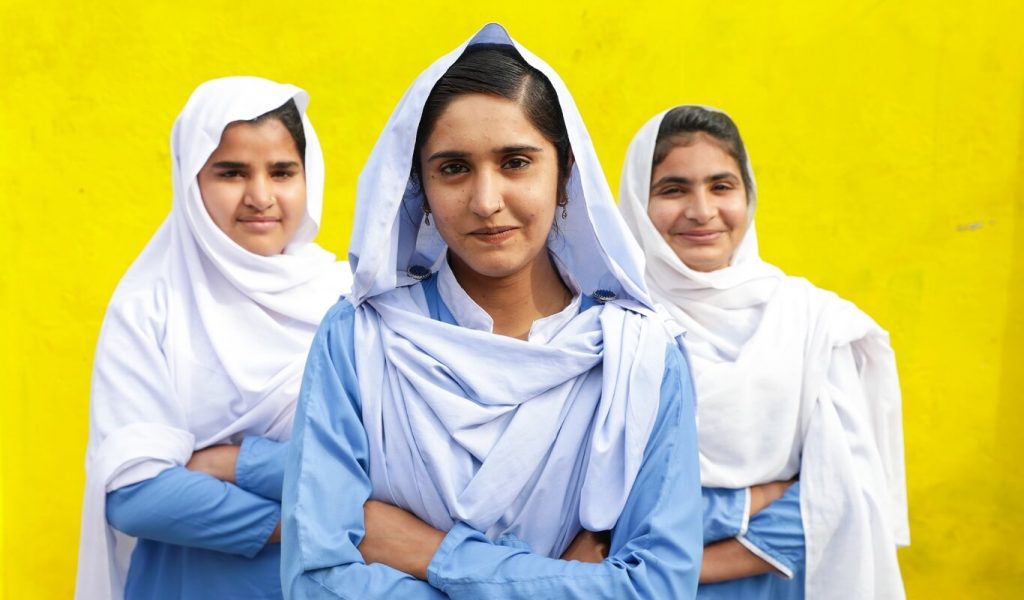 Three girls in white headscarves and blue dresses against a bright yellow wall cross their arms and smile at the camera