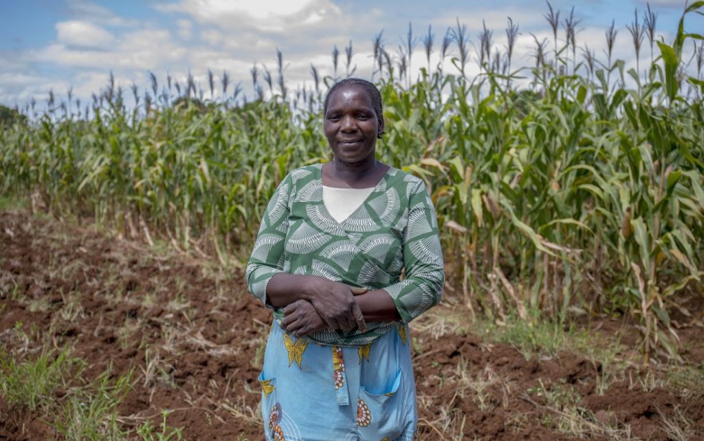 Woman in green floral shirt and blue patterned skirt with arms crossed smiling stands in front of crop field.