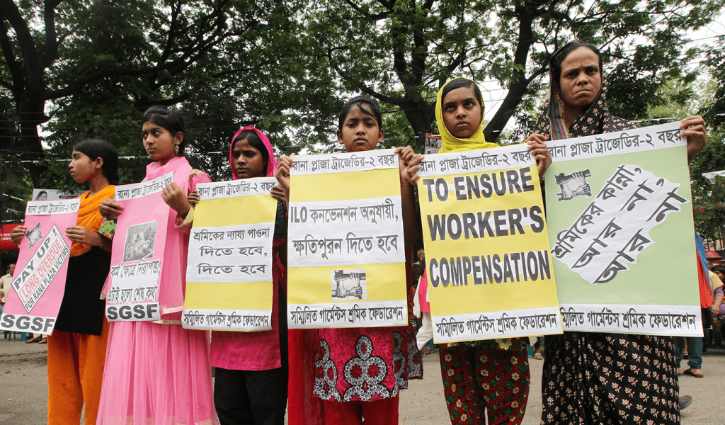 Six young women standing in a line holding pink, yellow and green protest signs in Bengali and English to compensate workers