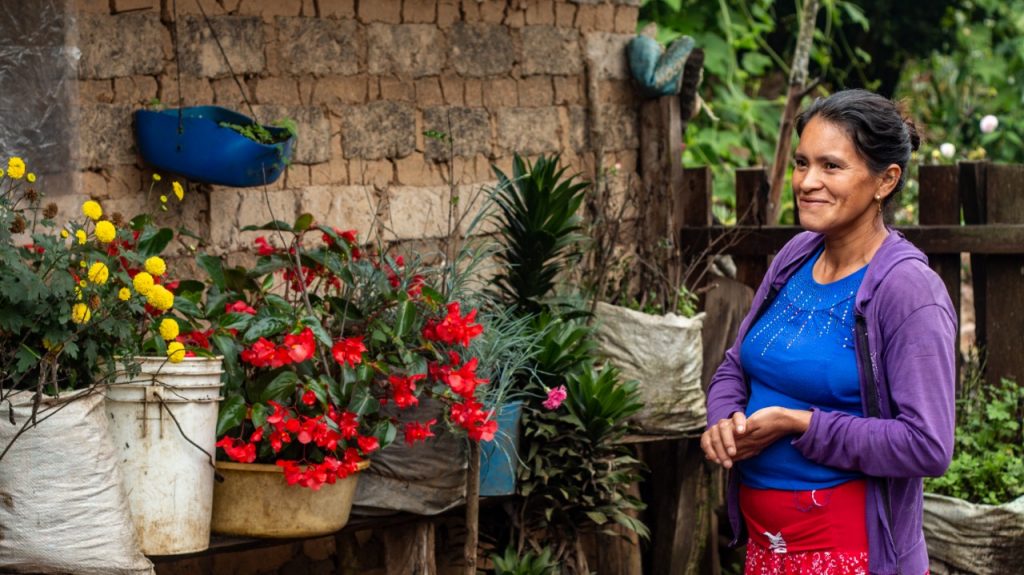 A woman wearing a red skirt, a blue blouse and a purple sweater stands to our right while smiling and looking to her pots of colourful flowers.