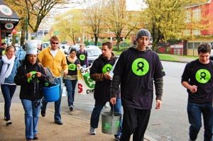 A Guatemalan woman and four young men all wearing black Oxfam shirts walk with buckets in their hands through a street in downtown Vancouver.