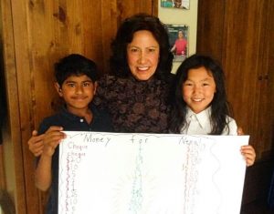 A Guatemalan woman stands smiling between a young South Asian boy and a young Asian girl in an elementary school classroom. They are holding a big piece of paper with a list of all the money they collected for an Oxfam emergency in Nepal.