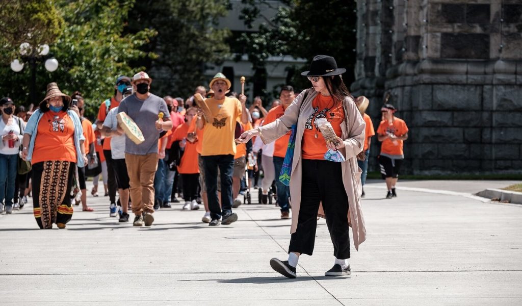 In the foreground, an Indigenous woman wearing a black brimmed hat, sunglasses, an orange tshirt, black pants and a long pink sweater scatters tobacco. In the background, dozens of Indigenous people and allies march while drumming and singing.