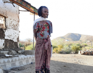 Woman in white scarf wearing grey shirt with red floral pattern and red skirt. Standing in front of eroded building and dry, grassy hill.