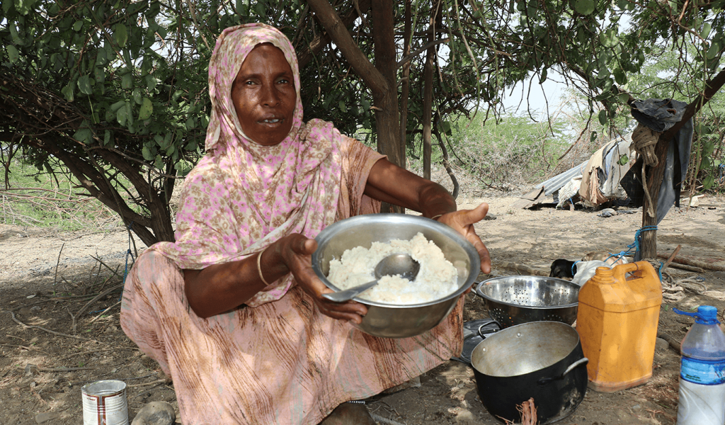 Elderly woman in pink, brown, floral hijab and pink dress sitting in front of trees on sandy ground. She is holding a metal pot with mashed potatoes in it. Sitting next to two other empty metal pots and a yellow jerry can.