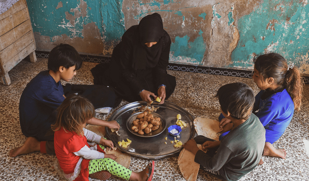 Woman in black hijab with her four children sitting in a circle around a metal pan that has pieces of potatoes divided amount each other. Woman is peeling the potatoes sitting in front of faded blue concrete wall and stone floor.