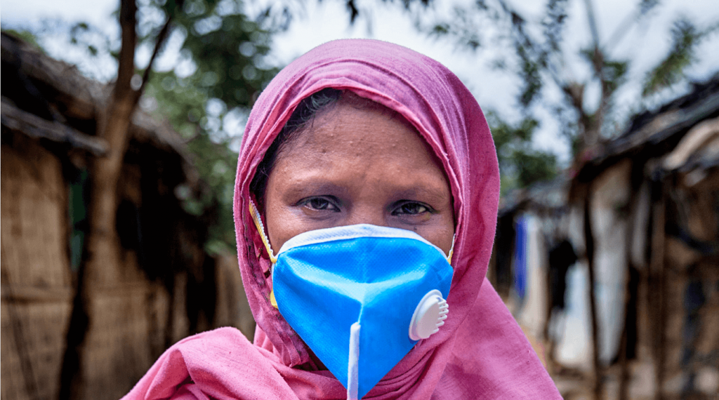 A brown-skinned and brown-eyed woman wearing a pink headscarf and a blue face mask looks directly at the camera. There are blurred trees and small huts in the background.