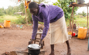 Woman in purple sweater and beige skirt leaning over lifting lid on metal pot. SHe is standing on dirt with grass and trees in the background. There are yellow jerry cans and chickens in the back.