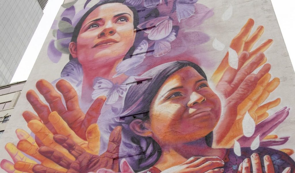 A multi-storey street art painting on the side of a building in an urban environment depicts a woman and child, both with brown skin. In the foreground, there are four sets of wrinkled hands of varying skin tones palms facing us with leaves and butterflies in the background.