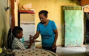 Elderly woman in blue shirt handing grandson in white pin stripe shirt a cup of water. They are standing in front of a yellow wall with a blue-green door to the right and a refridgerator to the left.
