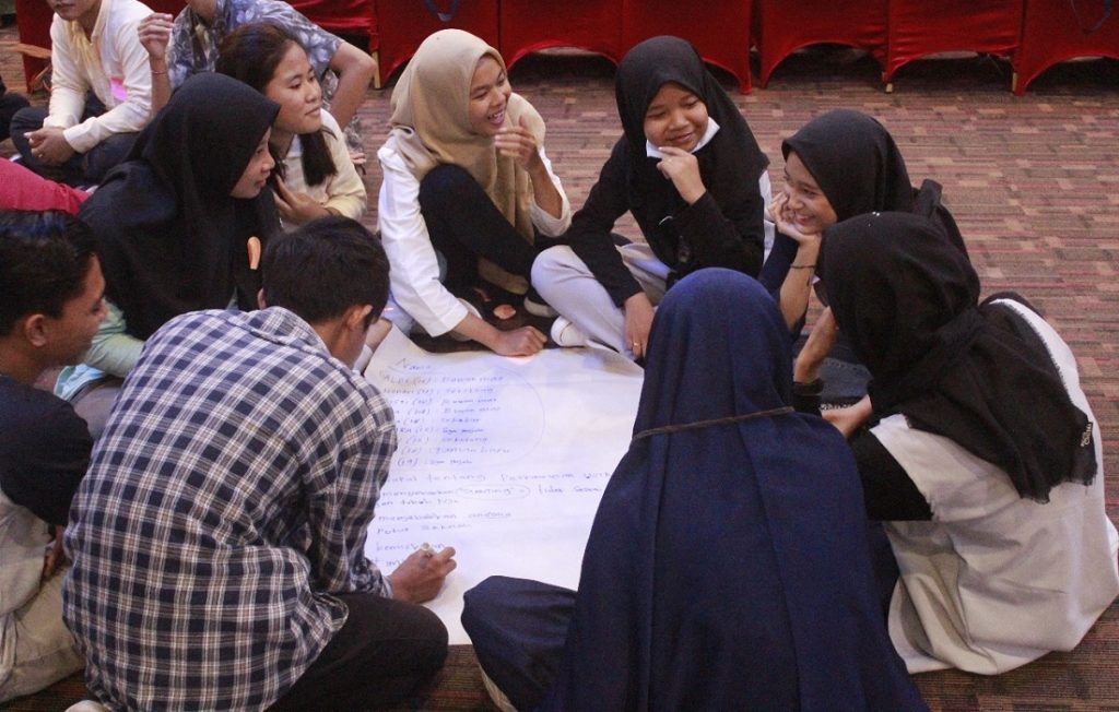 A group of 9 brown-skinned young women and young men gather in a circle around a large piece of chart paper. Some are talking, some are laughing, some are smiling and some are taking down notes. All the young women except for one are wearing head scarves