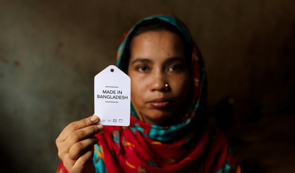 Woman wearing colourful hijab with red and turquoise pattern holding a white clothing tag with text that says "Made in Bangladesh". Standing in front of dark grey cement wall.