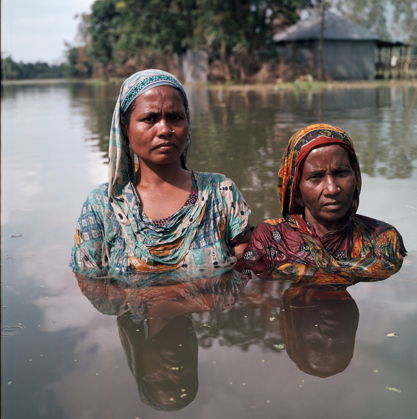 Two women standing chest deep in water. Woman on the left is slightly taller wearing a blue shayla and blue patterned dress. Woman on right is wearing an orange/red patterned shayla and dress.