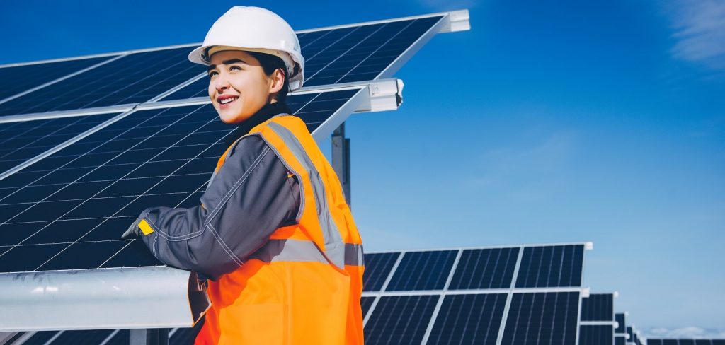 A young, light brown skinned woman in a hardhat and an orange reflective vest is inspecting solar panels on a solar farm