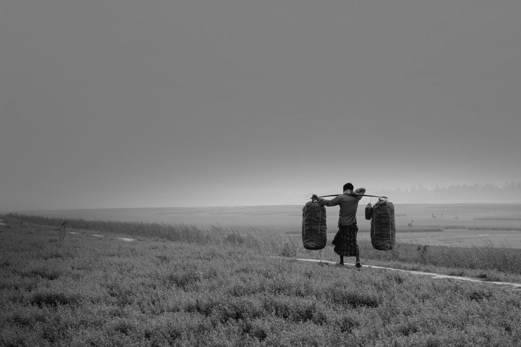 A Bangladesh person is walking along a dirt path through a field that is beside a river with a wooden pole that goes across their back and 2 large sacks on either end. The baskets are carrying their crop. It's a dark, dreary day.