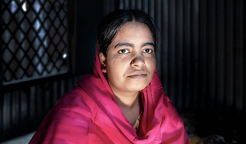 Portrait photo of person draped in pink head/body scarf making eye contact in a dimly lit room.