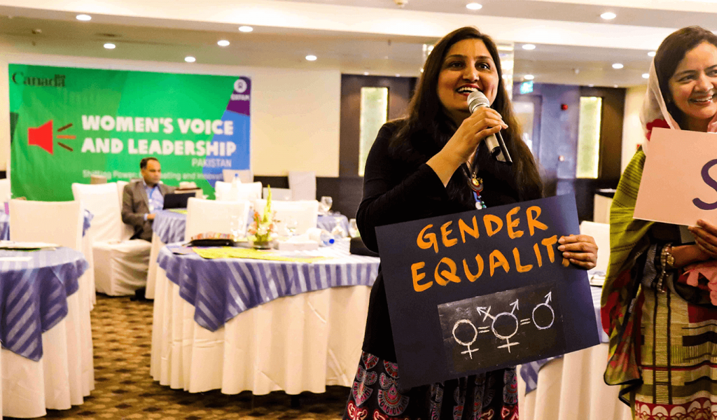 Person holding microphone with her left hand and a poster with her right hand. The poster is dark purple with the words "Gender Equality" written in yellow and the symbol for female equalling the symbol for transgender equalling the symbol for man. She is standing in a conference room with a poster in the background that says "Women's Voice and Leadership Pakistan" with the Government of Canada logo to the top left and Oxfam to the top right.