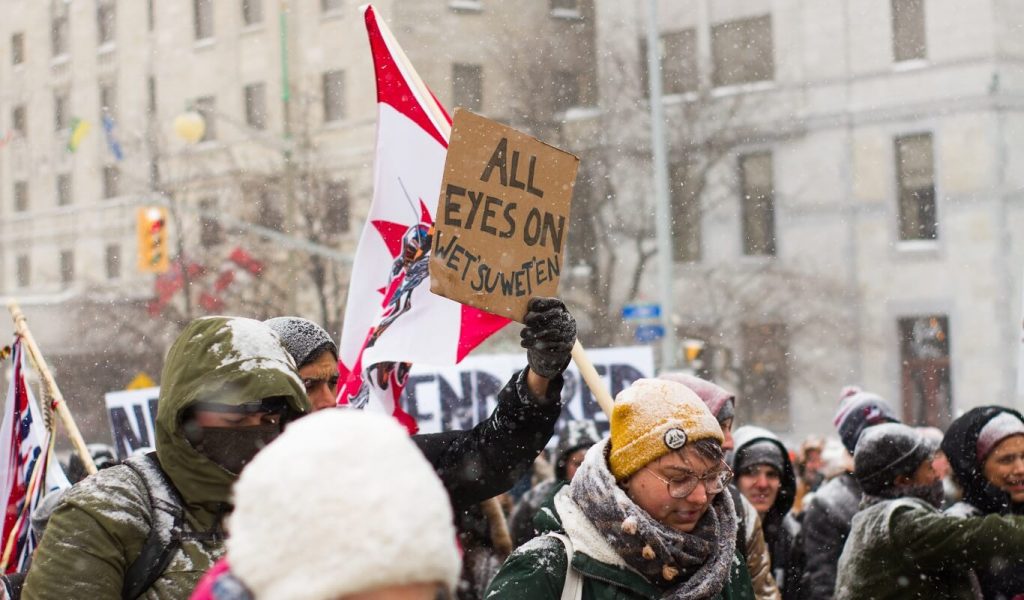 A group of people of various genders, ages and skin tones are bundled up against the cold as they march together in a daytime protest down a snowy street in downtown Ottawa, Canada. Many people are carrying protest signs. but only one sign, which is being held up in the the middle of the photograph, is visible. It says All eyes on Wet'suwet'en. Behind this sign, there is a Canadian flag.
