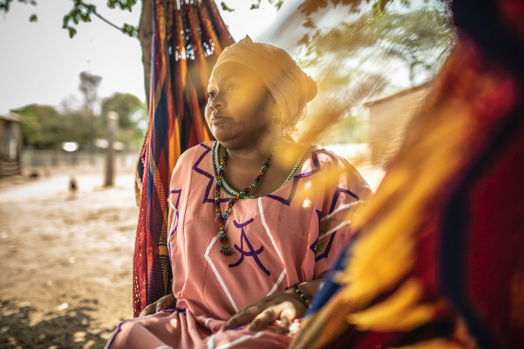 A black woman in colourful African clothing sits on the ground in a rural area and looks off into the distance.