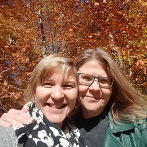 Two caucasian middle aged women stand cheek to cheek smiling outside in front of trees with yellow and orange leaves