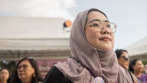 A young woman wearing a brown headscarf and beige glasses has a pin on her scarf that reads, I am a girl defender. She is standing in a group of women and looking upwards in an optimistic stance.