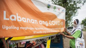 A black woman wearing a green t-shirt and a white and black headscarf hangs an orange banner that reads Labanan and COVID-19.