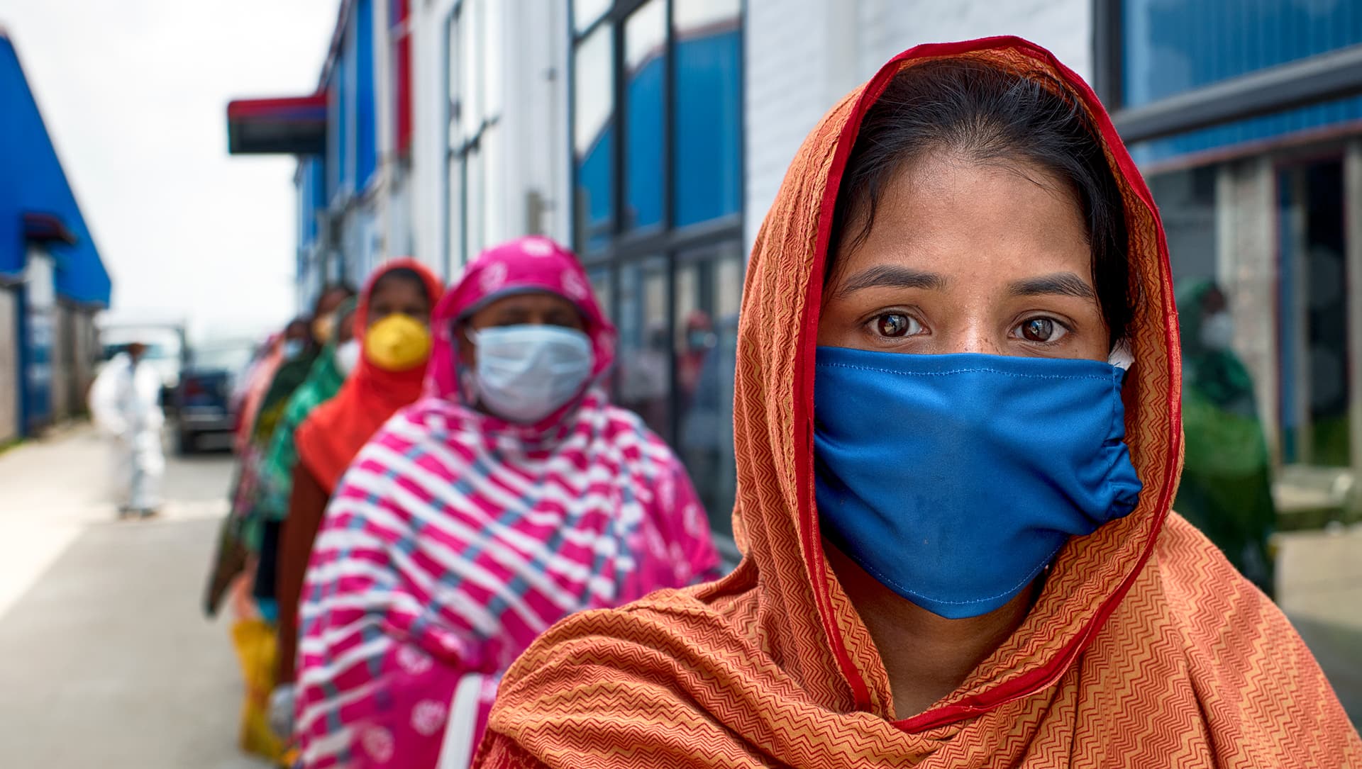A woman wearing a red and orange headscarf and a blue face looks into the camera, a line of other women stand behind her
