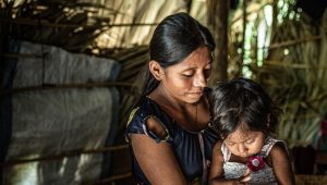 A young woman and her toddler daughter are sitting in a dimply lit thatch structure and looking downward