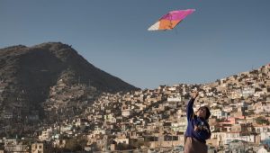 A young boy flies a pink and orange kit in rural Afghanistan, with a village of houses on a hill behind him.