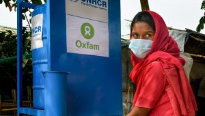 A woman wearing a red headscarf and blue medical face mask is washing her hands at a blue hand washing station that has the Oxfam logo displayed on it