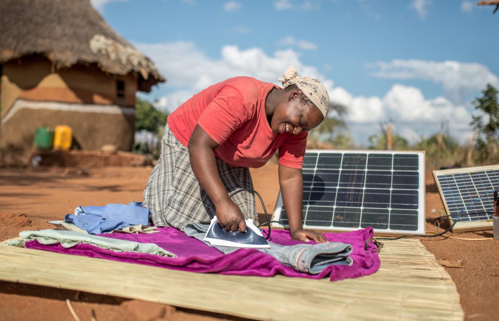 A woman is outside her home, smiling, while on her knees, next to a couple of solar panels that power the iron she's using to iron a pair of blue jeans.