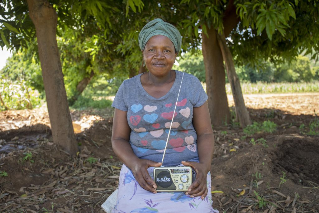 A woman, in the centre of the image, sits among some trees. On her lap she holds a radio while staring directly at the person who shot the image.