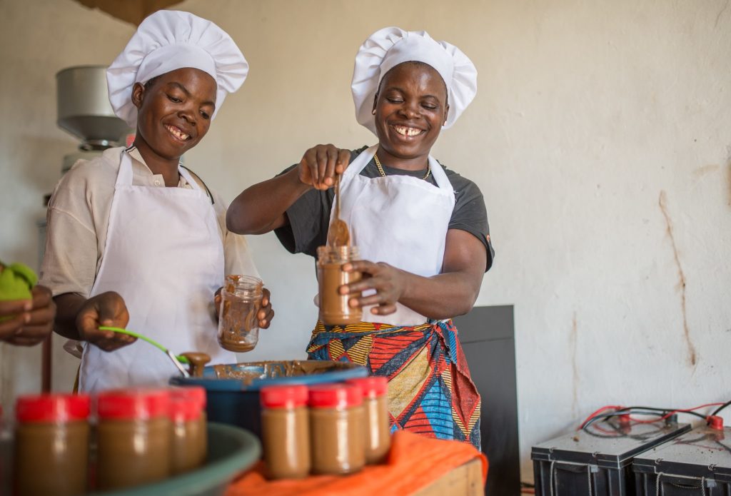 Two smiling women wearing white hairnets and aprons, bottle crystal jars with peanut butter.