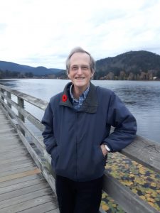 An older Caucasian man dressed in a blue jacket and black pants stands smiling on a dock by the water with hills in the background.