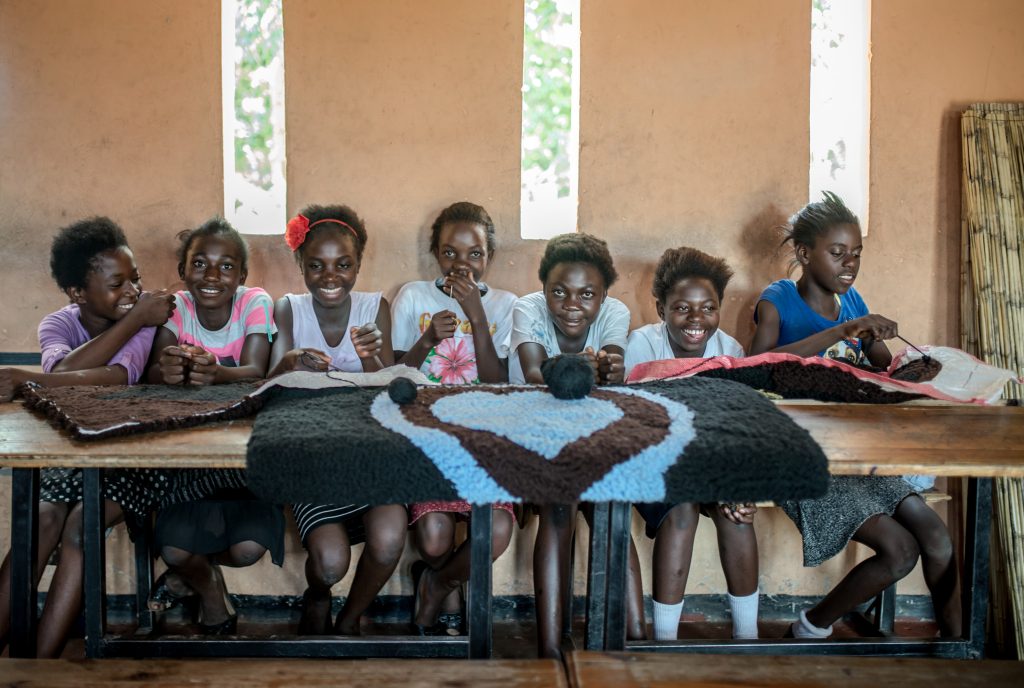 Inside a room, seven young black girls sit smiling at a long wooden table with handmade rugs across the table.