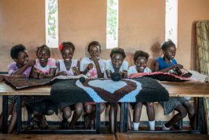 Inside a room, seven young black girls sit smiling at a long wooden table with handmade rugs across the table.