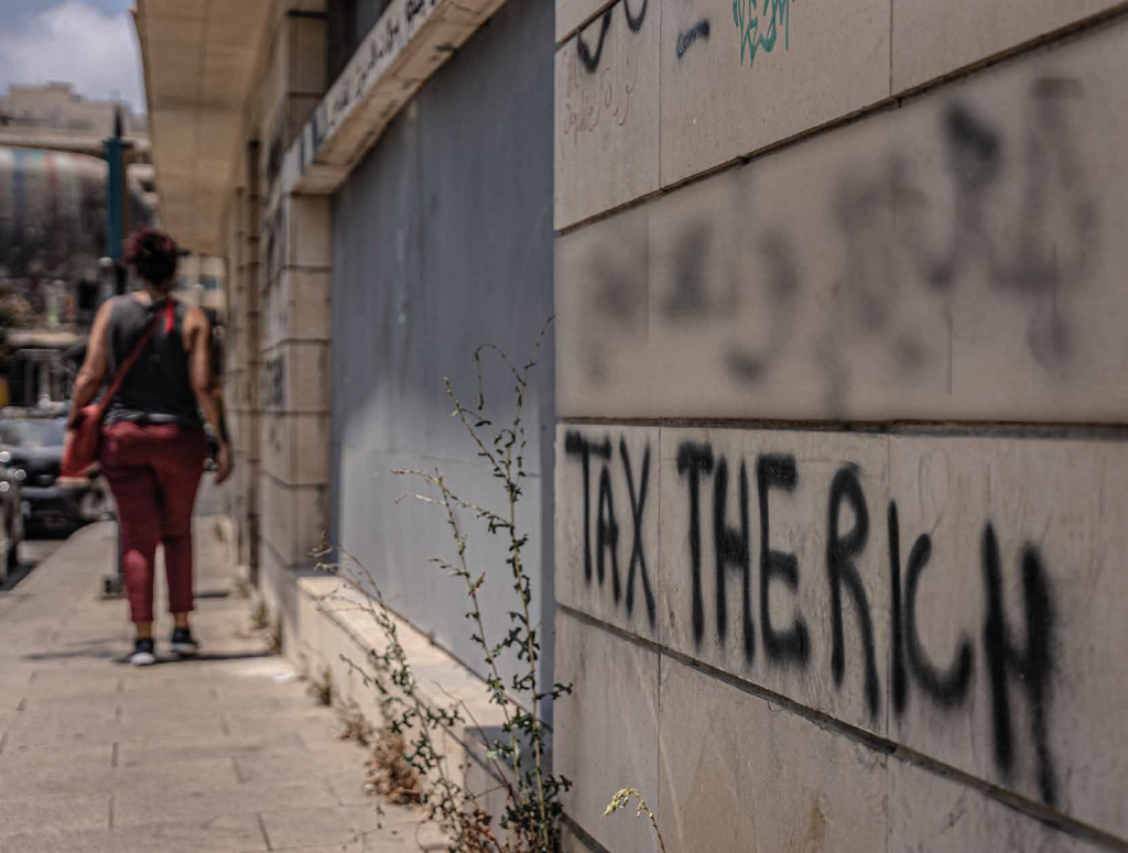 In this candid shot, we see a wall of an urban building on the right marked with black graffiti that reads tax the rich. To the left of the building, there is a dark-skinned person walking down the sidewalk facing the other direction in a black sleeveless shirt, red capris pants, black sneakers and a red satchel.
