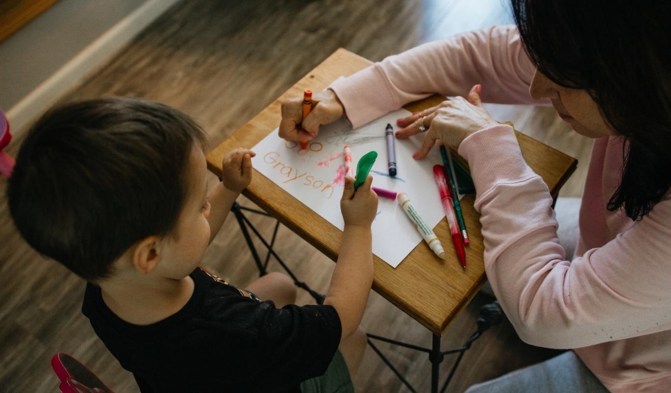 A young light-skintoned child with short, dark hair, wearing a black t-shirt and olive green shorts sits at a small rectangular desk. On the desk is a piece of paper with the name Grayson marked on it along with drawings and doodles, along with several crayons of various shades. Crouched down next to the desk writing on the paper with the child is a medium-light skintoned middle-aged adult with dark, shoulder-length hair. She is wearing a pale pink hoodie and leggings.