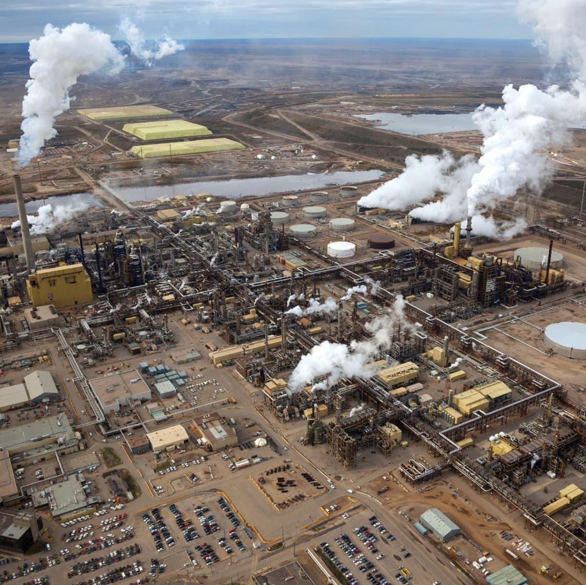 An overhead view of an oil refinery from the sky. There is a network of buildings, industrial-sized vats and tall smokestacks emitting smoke and other pollution. In the distance, we see a river.