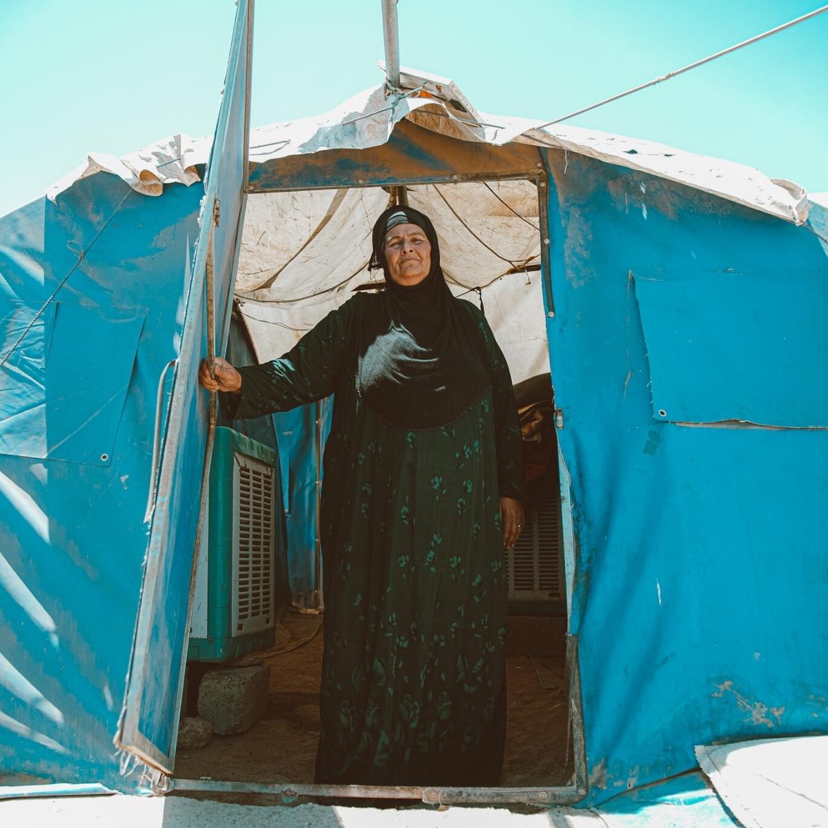 An elderly light-skinned woman stands in the doorway of her family's light blue tent. The woman is wearing a long-sleeved ankle-length dark green patterned dress and a black headscarf. Behind her, the sky is clear and bright.