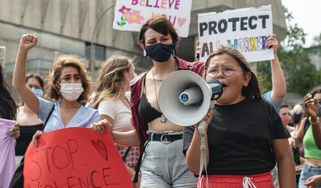 A crowd of feminine-presenting people of varying ages is gathered outdoors with homemade protest signs that say: Protect all workers, I believe you and Stop violence against women. On the bottom right of the image, there is a child of approximately 10 years old speaking on a bullhorn while visibly upset, face contorted and tears in her eyes.