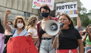A crowd of feminine-presenting people of varying ages is gathered outdoors with homemade protest signs that say: Protect all workers, I believe you and Stop violence against women. On the bottom right of the image, there is a child of approximately 10 years old speaking on a bullhorn while visibly upset, face contorted and tears in her eyes.