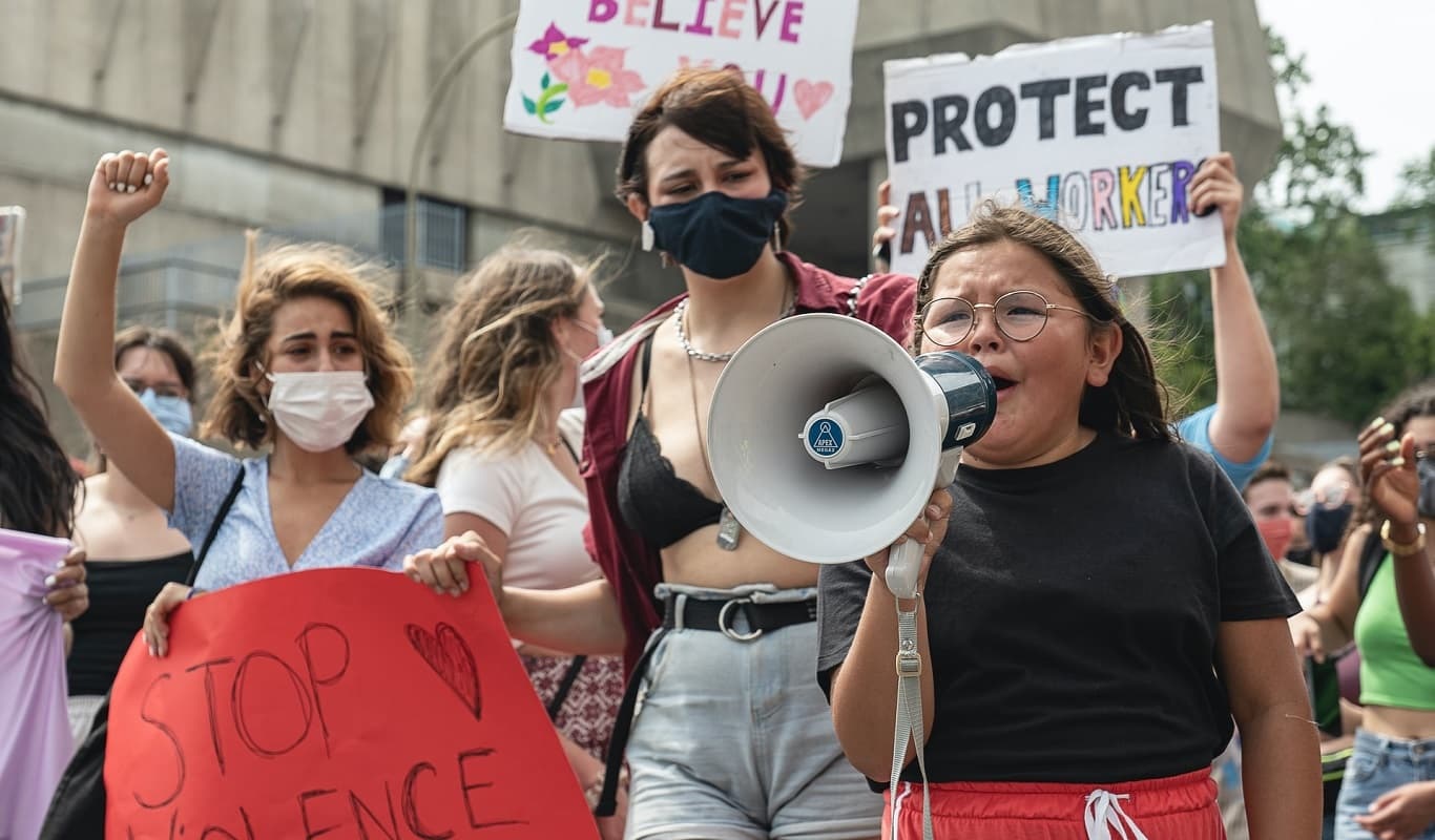 A crowd of feminine-presenting people of varying ages is gathered outdoors with homemade protest signs that say: Protect all workers, I believe you and Stop violence against women. On the bottom right of the image, there is a child of approximately 10 years old speaking on a bullhorn while visibly upset, face contorted and tears in her eyes.