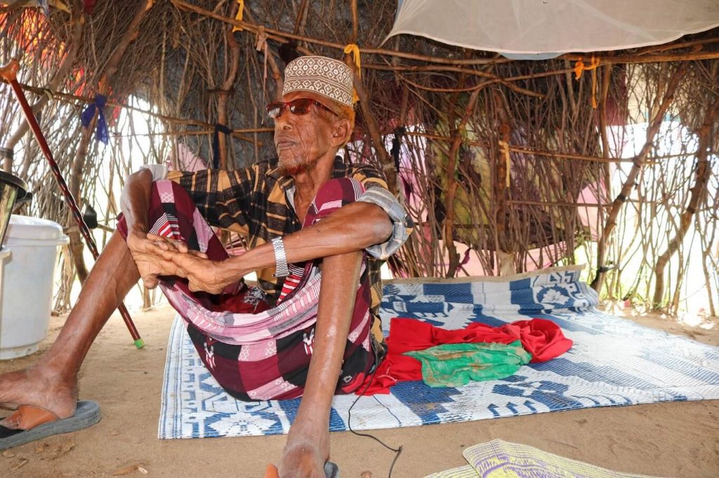 An older, dark skinned man sits on a blue and white plastic blanket under a hut made of long, thin sticks. He is wearing sun glasses, a sliver watch, beige flip flops and a black and orange checked shirt with the sleeves rolled up to his elbows and a wine and black traditional wrap skirt that goes just past his knees. He's sitting on the blanket with his knees bent and arms rest on top of his knees.