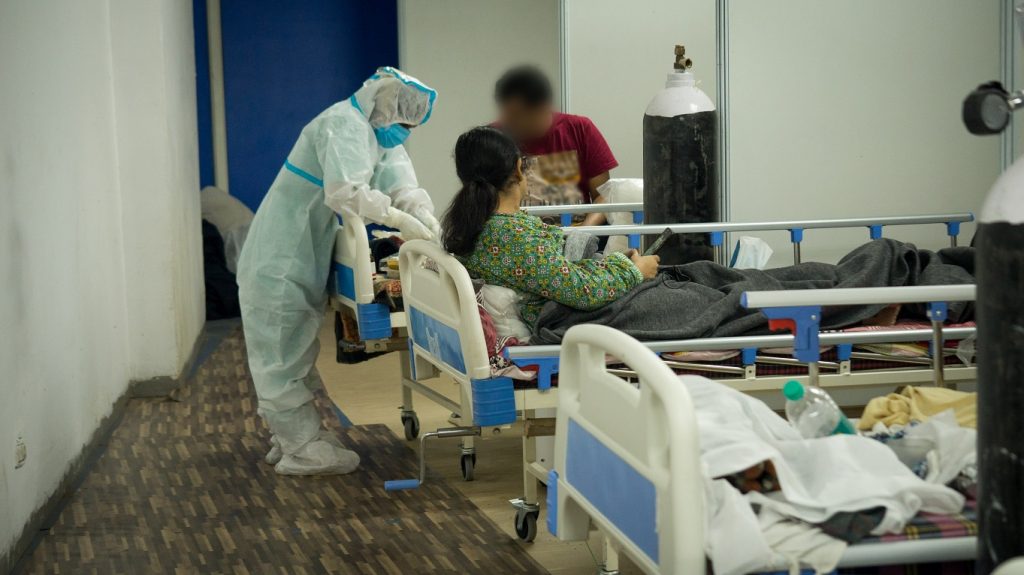 A nurse dressed in a white paper suit is bending over a bed looking after a patient. There are 3 bed ends with white railings and light blue panels. There is a brown woman with long dark hair in the middle bed. She is sitting up and looking towards the nurse. In the background is a man with a red shirt and an oxygen tank.