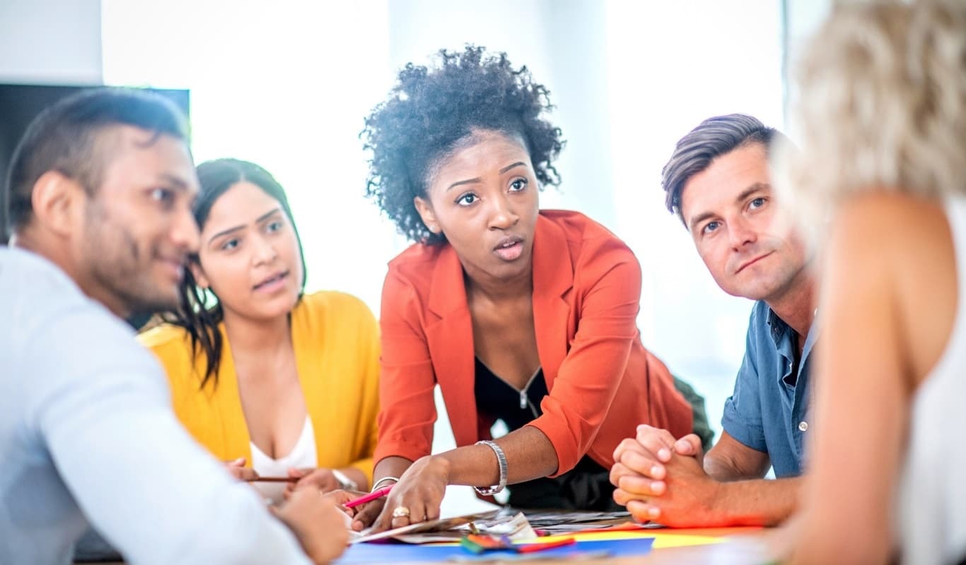 A group of five early to middle-aged adults ofa various ages and skin tones are gathered around a table in a working environment. The focal point of the picture is a young dark-skinned woman in a black tank top and an orange blazer leans across the table pointing at a document.